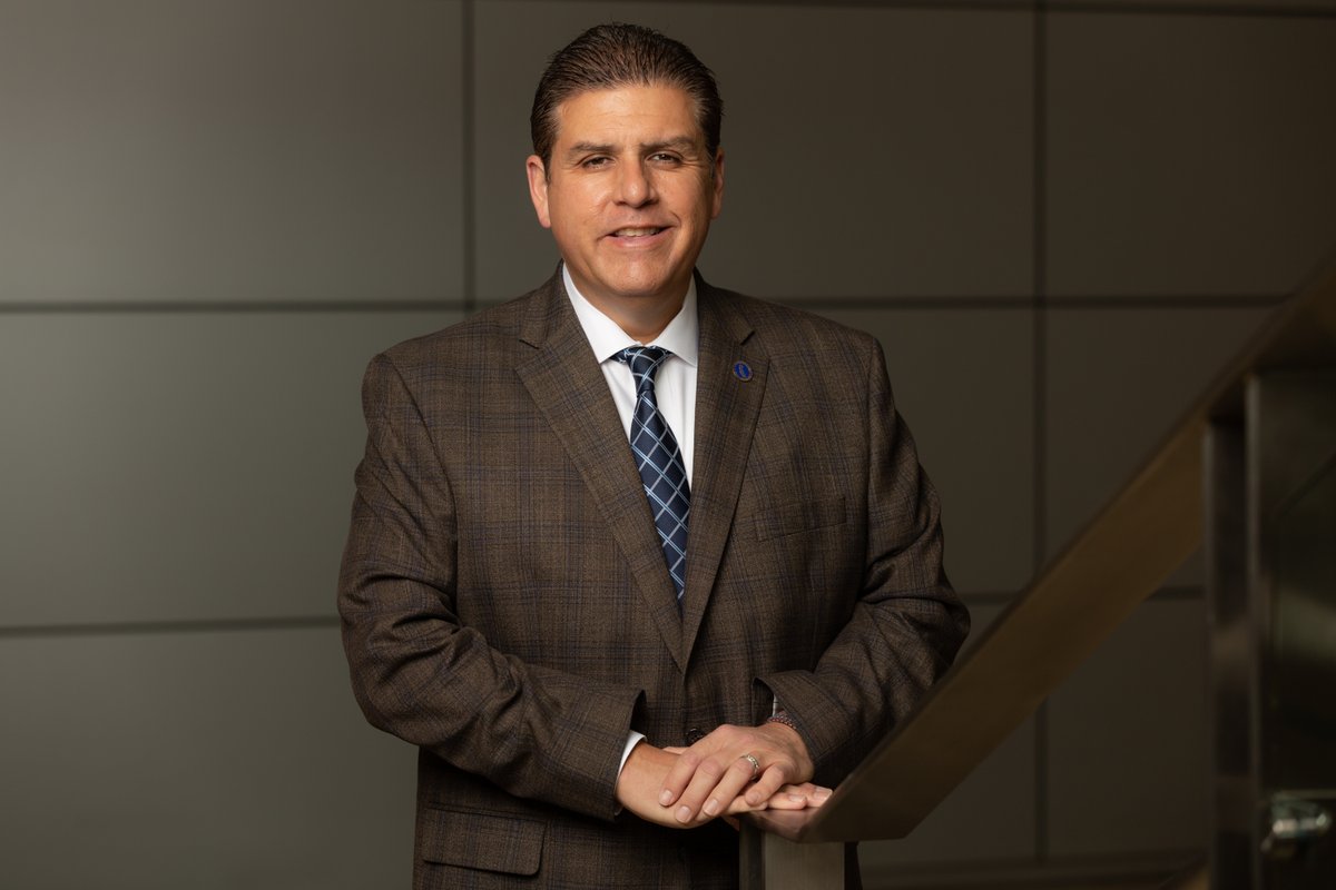 The California State University President, Joseph I. Castro standing at the bottom of a stairwell. Castro is in a brown suit with a grey wall behind him.