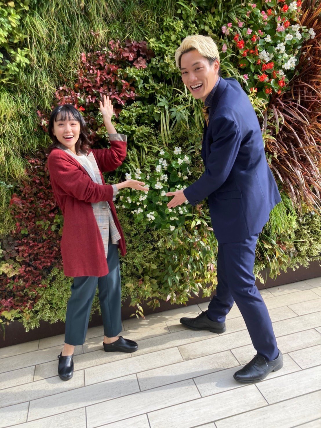 ５話のオフショット写真。場所は街中の通り。満面の笑みのイズミとシシオが、花や植物が敷き詰められた壁の前で、並んで立っている。２人とも花のほうに手を向けている。シシオの服装は、コンのスーツ、コンのシャツ、ゴールドと茶色のストライプのネクタイ。イズミの服装は、白のブラウスにワインレッドのカーディガン、グレーのパンツスタイル。