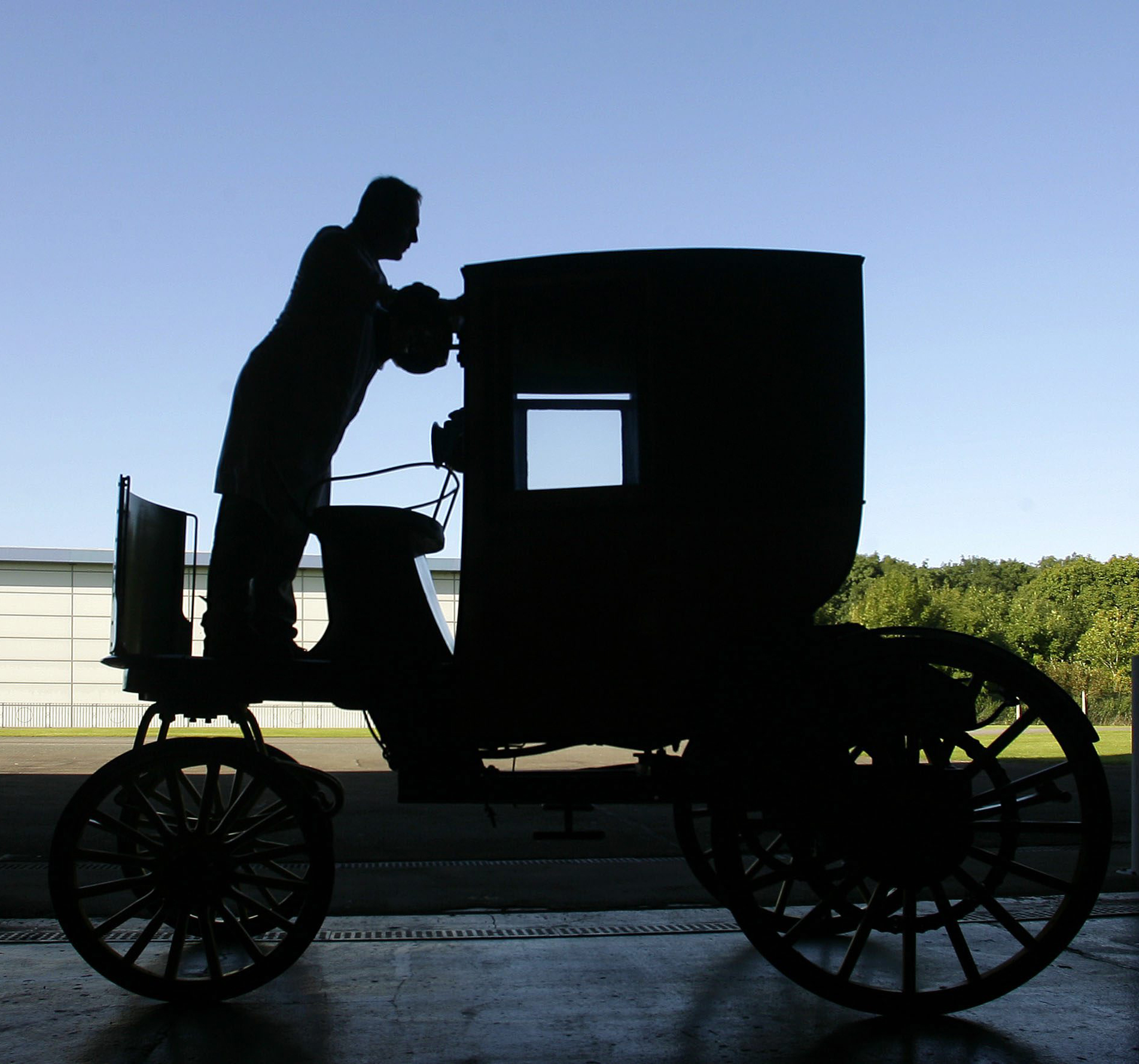 A black silhouette of a man standing next to an antique carriage, against a sunny blue sky. It can be seen that the carriage has 4 large spoked wheels and a small cab, with seat at the front for the driver. It is slightly taller than a person.