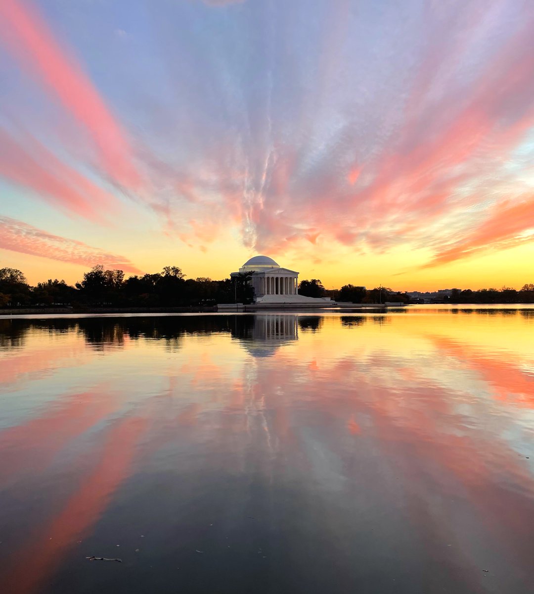The white domed Jefferson Memorial stands at the other side of a still pond under a colorful sunset sky.
