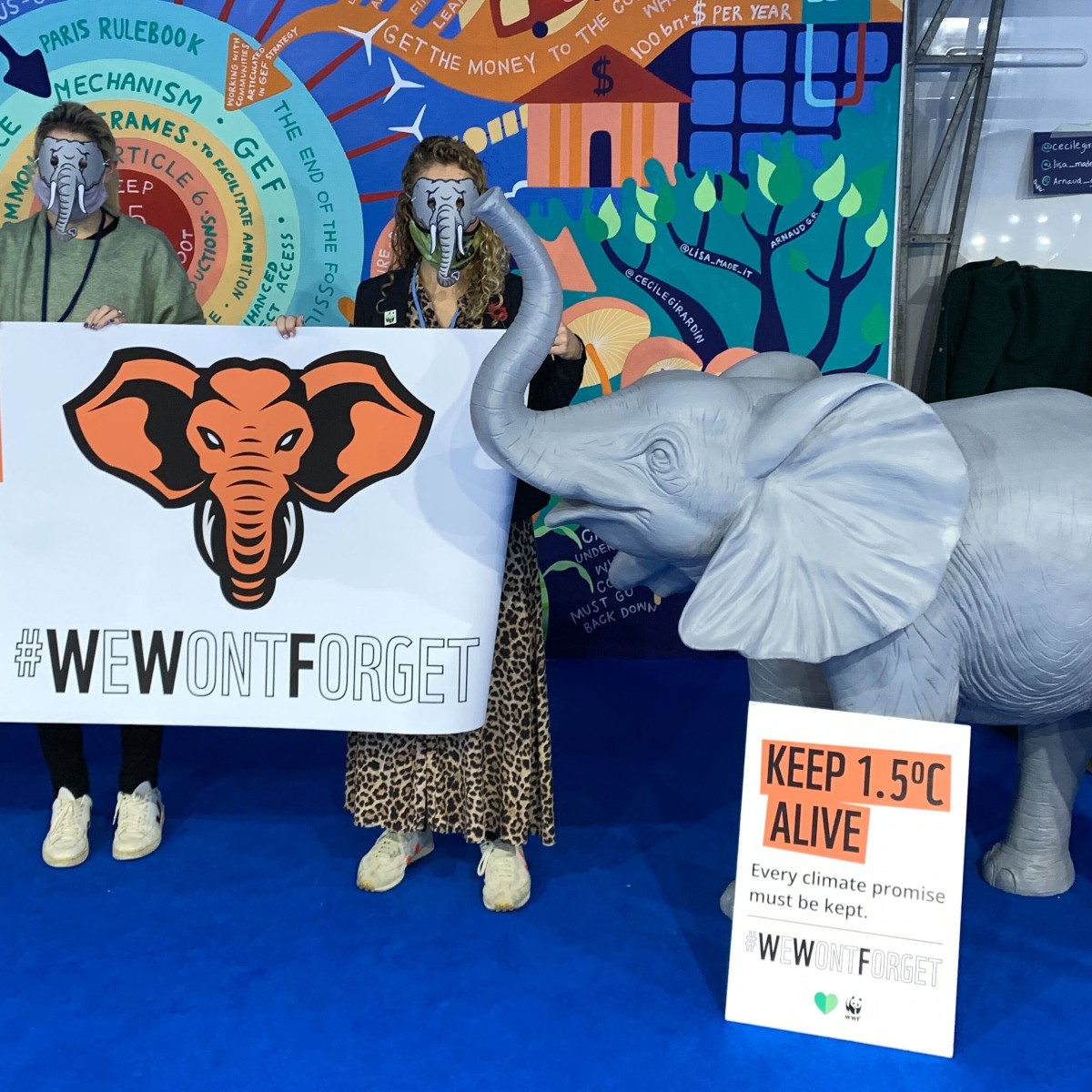 A life-size baby elephant model next to people holding a large banner that reads 'We Won't Forget'