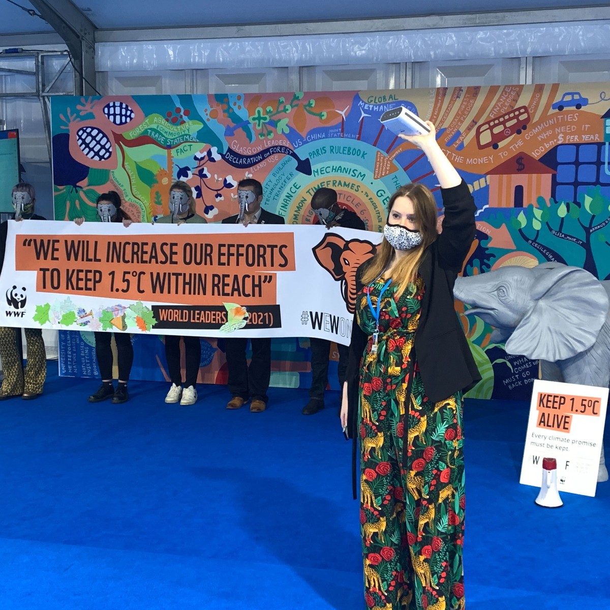 A woman stands in front of a protest banner holding a speaker in the air
