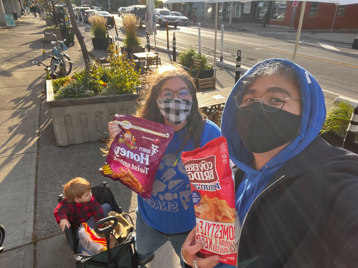 Terri, a caucasian female presenting person with a blue hoodie that has a snack graphic is next to her child in a wagon. Terri is holding up a korean sweet snack next to Carl, a non-binary asian person, wearing a blue hoodie and holding a bag of ketchup chips. They are both wearing masks and looking straight at the camera. 