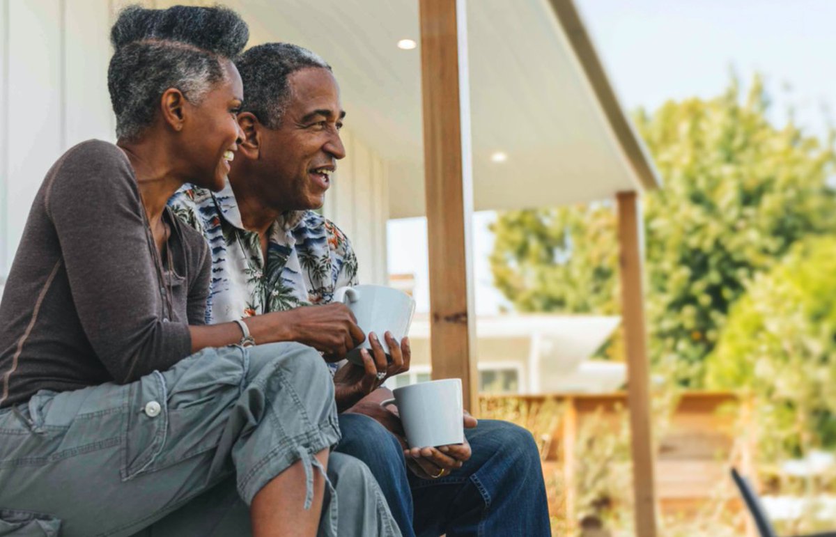 Couple sitting on the porch of a home holding mugs and looking out over the yard.