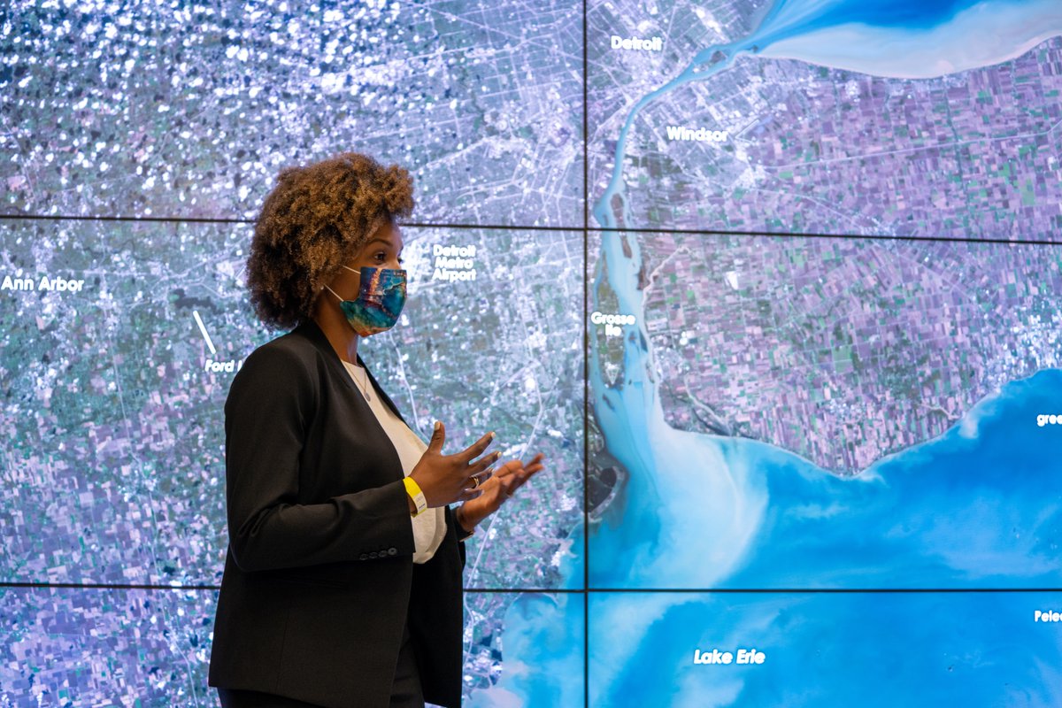 Dr. Lola Fatoyinbo stands in front of several TV screens showing a satellite image of Detroit. She is talking with her hands