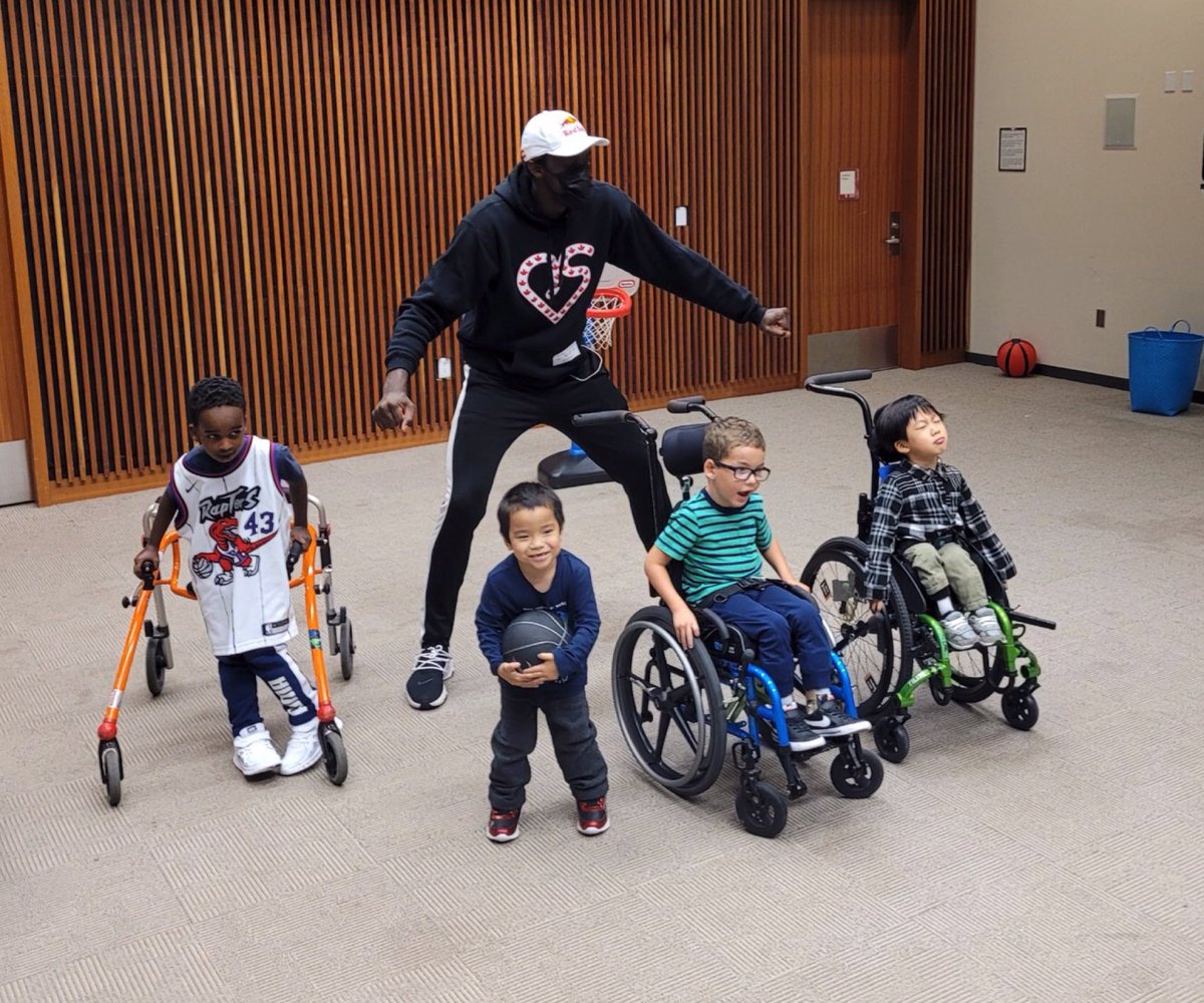 Pascal Siakam stands behind four children with his arms outstretched. One child is wearing a Raptors jersey and is using a walker. The next child is standing and holding a basketball. Another child wears a striped shirt and glasses and is using a blue wheelchair. The final child is wearing a plaid shirt and using a green wheelchair.