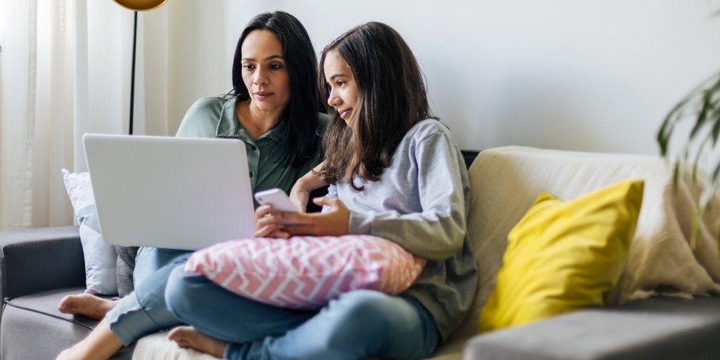 A mother and daughter, both dark skinned with long black hair, wearing a green and grey hoodie respectively. They are sitting on a grey couch looking directly at the screen of a silver laptop. There are yellow and pink and white stripped pillows on the couch. They appear to be in a living room.