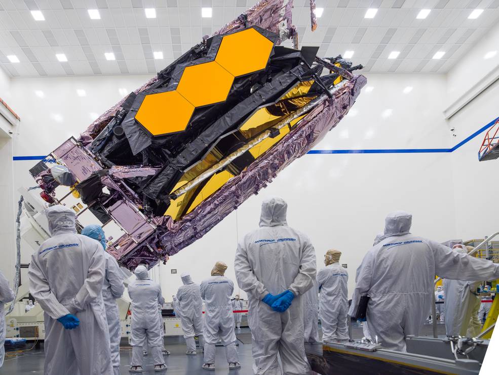 NASA's James Webb Space Telescope is tilted by cables as it is lifted and prepared for transport to French Guiana inside the Northrop Grumman cleanroom. Several technicians in white cleanroom suits observe the telescope, their heads turned away from the camera.