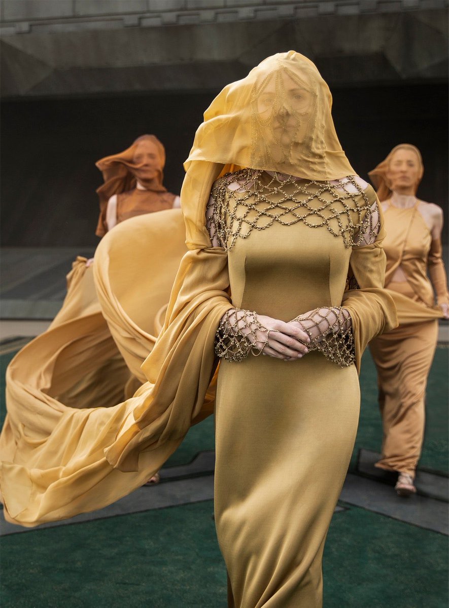 Lady Jessica (Rebecca Ferguson) in DUNE (2021) walking across the airfield. It's golden yellow, includes a sheer veil over her face, and an elaborate beaded head cap. © Warner Brothers