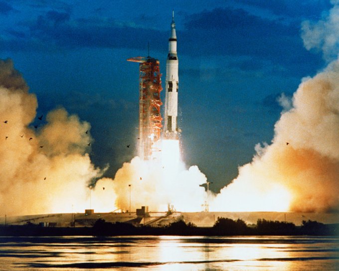 Photograph of Apollo 4 launching. A large, white and black rocket lifts off and smoke and fire billow from below. 