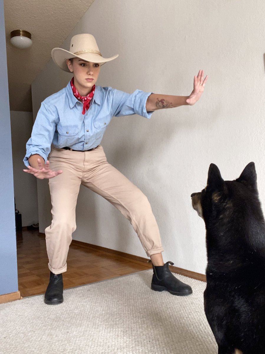 A photo of Ellie dressed as Dr Alan Grant from Jurassic Park, in a light blue denim shirt with the sleeves rolled up, a patterned red bandana tied around her neck, a tan western style hat, tan pants, a black belt and black chelsea style ankle boots. In front of her is her dog, Kubo, a Black and Tan shiba, sitting facing away from the camera and towards Ellie