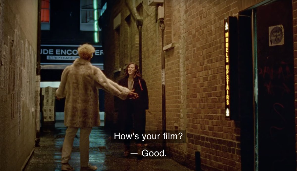 Two friends in an alleyway. The man on the left is wearing a fur coat and has his arms out. And the woman on the right is wearing a black coat. Caption: "How's your film?" "—Good."