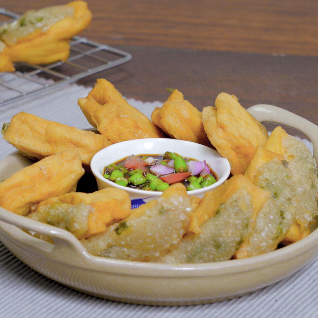 A bowl of golden fried wontons/pastries arranged around a small dish of soy-based dipping sauce containing edamame beans and onions.