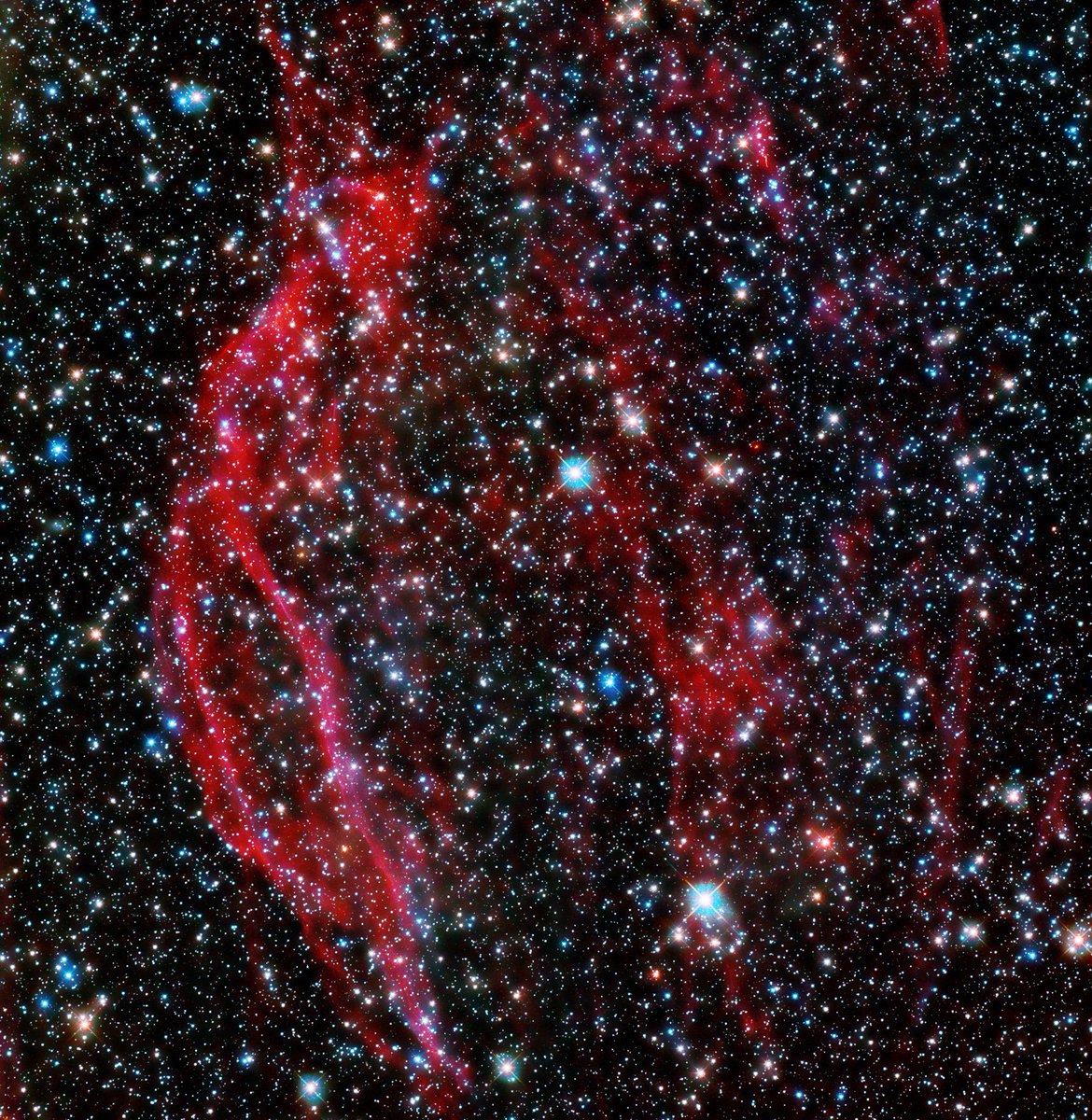 Millions of stars twinkling on the black backdrop of space with red cosmic ribbons of gas flowing from top to bottom of the scene. 