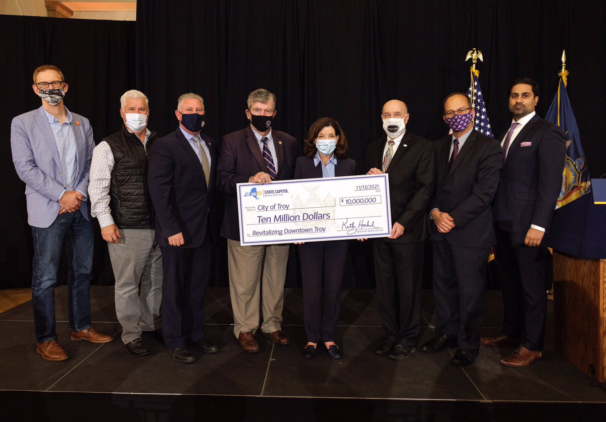 Group of 8 people standing in line holding a 10 million dollar check to the city of Troy
