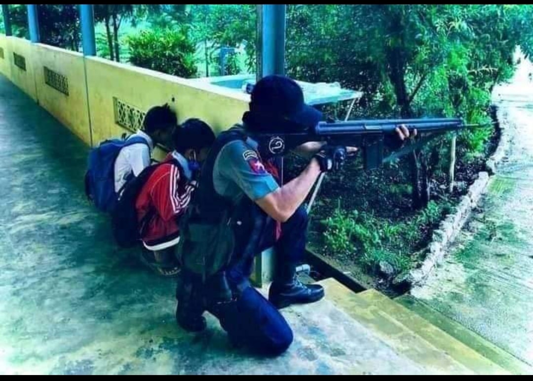 Khinmau11618908's tweet photo. Myanmar School children are under attacked by gun  fire of PDF terrorists. The image show a police man shooting back at the PDF terrorist while two small children hiding behind the wall of a school corridor. Another image is a Buddhist monk murdered by PDF. https://t.co/0u5DGdwWID
