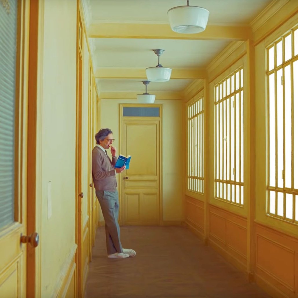 Christoph Waltz as Boris, wearing mauve cardigan and trousers. He leans against wall in corridor painted yellow in The French Dispatch.