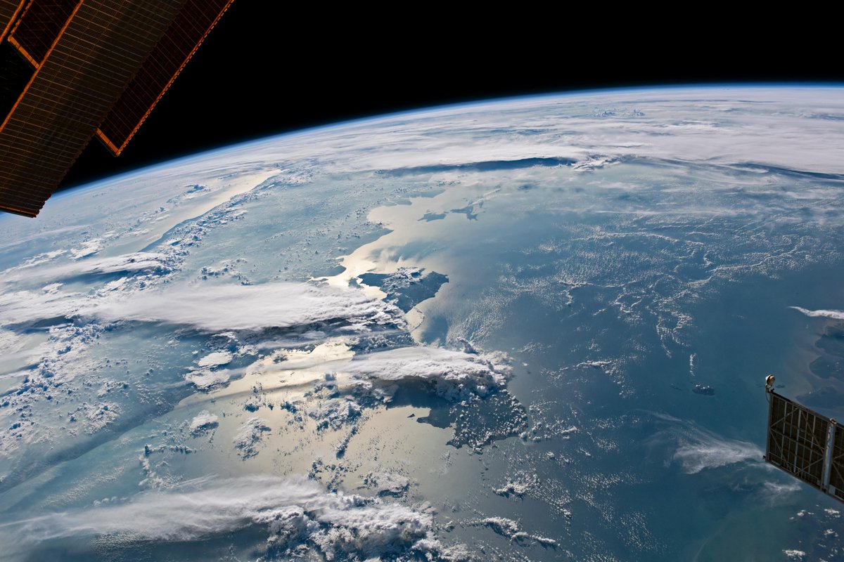 Photograph of a light-blue Earth taken from space, with black looming above a glowing blue horizon line. Long, horizontal strips of clouds rest over multiple islands. Two ISS solar panels peek out in the corners of the image.