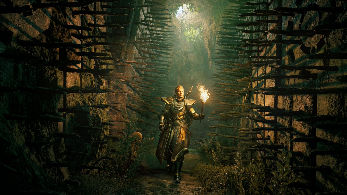 Eivor walks towards the camera with a torch in her left hand within a cave system. The walls are close, covered in spikes - a trap waiting to be sprung. A small beam of light breaks through the ceiling, illuminating a faint green mist.
