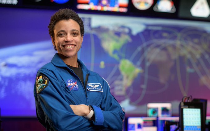 African American astronaut in blue flight suit, with arms crossed. She's smiling and the Houston mission control center's large screen is out of focus in the background.