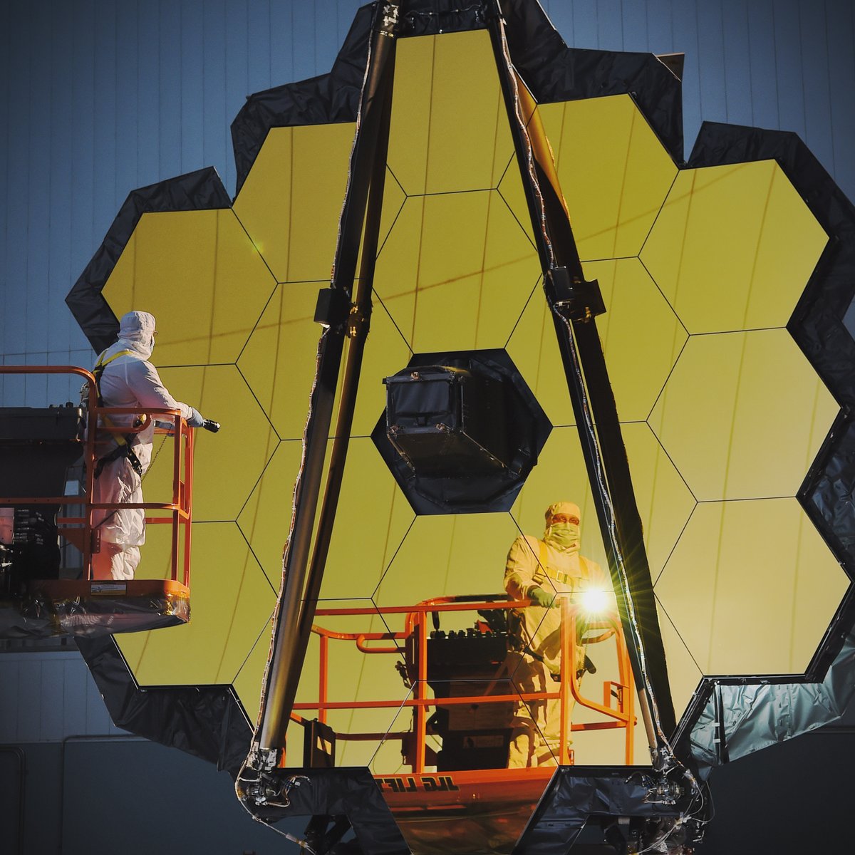 The James Webb Space Telescope's primary mirror is lit up in the dark. A single technician stands on an orange crane, holding a flashlight to the mirror.