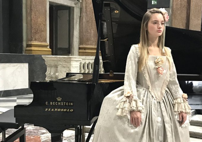 A woman (Fedora Florian) wearing a costume curated by Pasquale Napolitano, stands before a grand piano. 