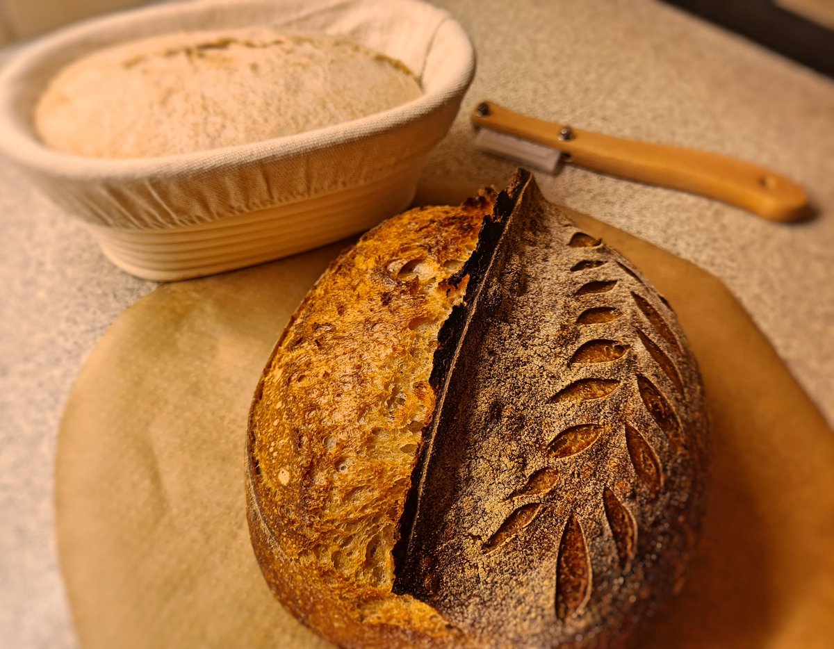 A finished loaf of sourdough bread by a banneton of dough and a lam