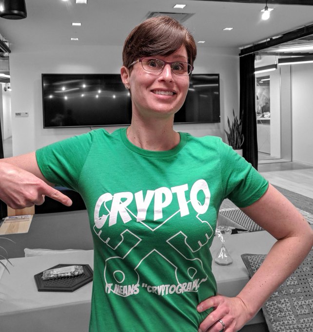 Lea in an extremely grey office area wearing a green tee that says "CRYPTO it means cryptography" on it with crossed keys behind.