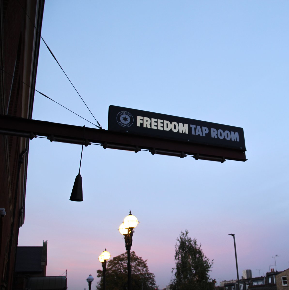 An image of a sign extended from a red brick building against a dusky sunset and some street lamps. The black sign reads 'FREEDOM BREWERY'.