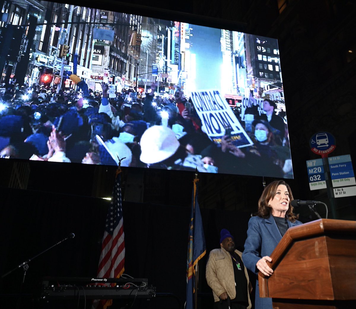 Governor Hochul (far right) stands at podium in front of large screen