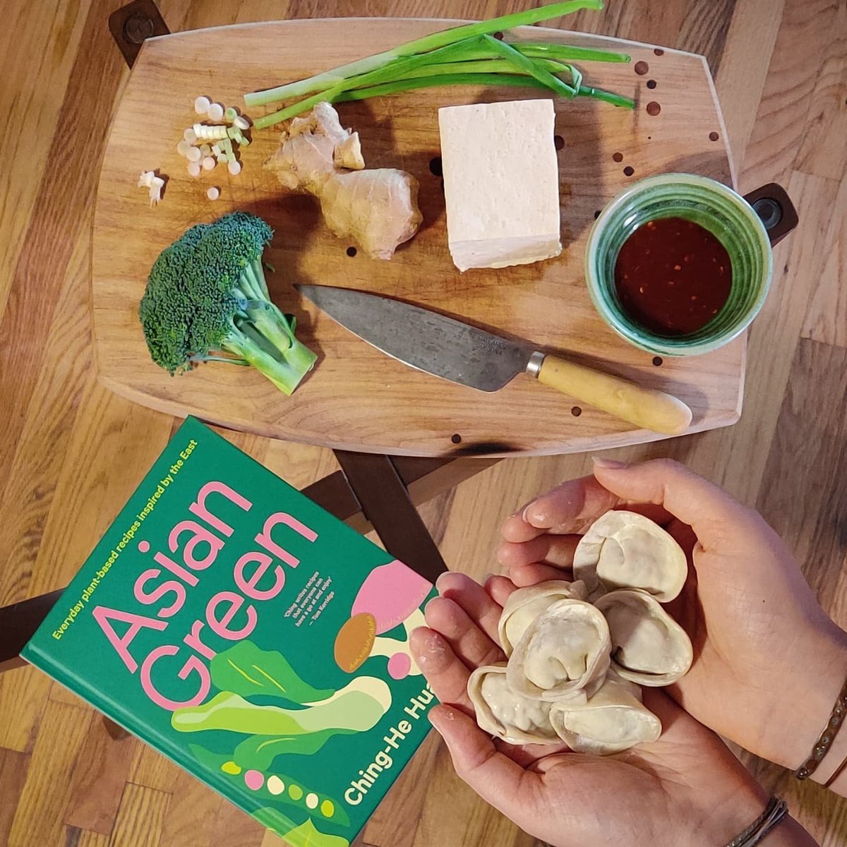A pile of uncooked dumplings are cradled in a pair of hands beside a cookbook titled "Asian Green." In the background, a cutting board is visible with tofu, garlic, spring onions, broccoli, chili sauce, and a knife.