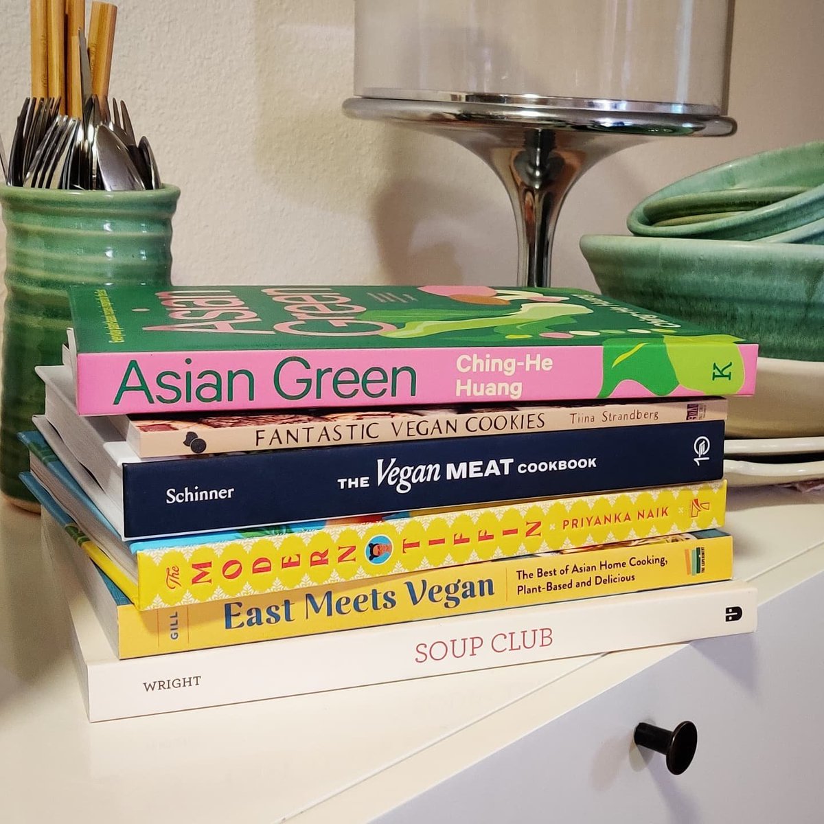 A stack of cookbooks sit on a kitchen counter. Their titles are "Asian Green," "Fantastic Vegan Cookies," "The Vegan Meat Cookbook," "The Modern Tiffan," "East Meets Vegan," and "Soup Club."
