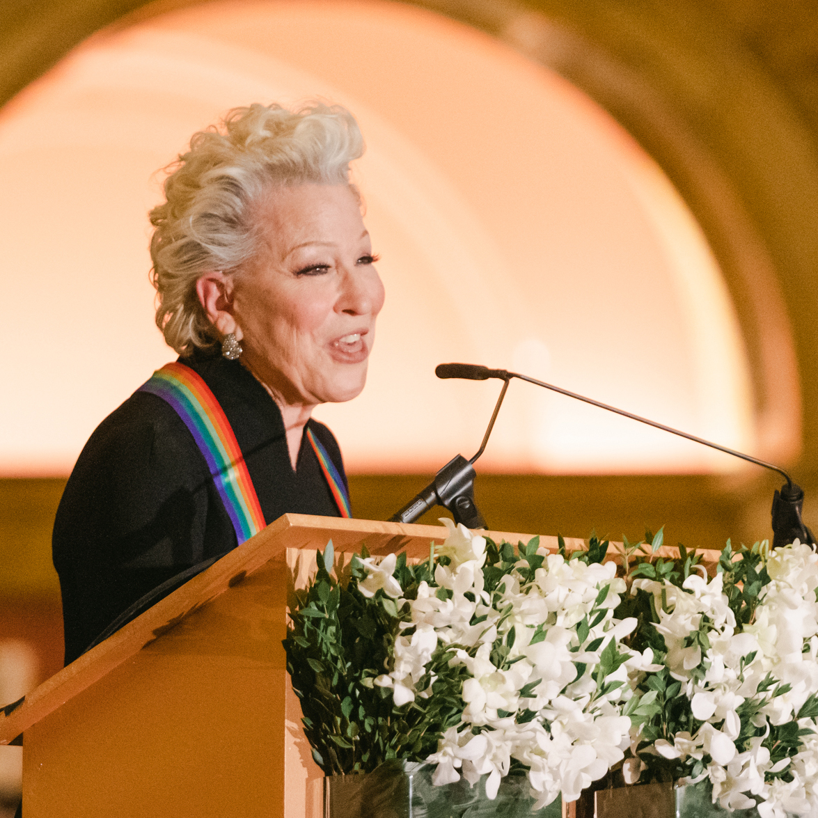 Bette Midler stands at a podium smiling & speaking into a microphone, wearing a rainbow Kennedy Center Honors medallion and a black top.
