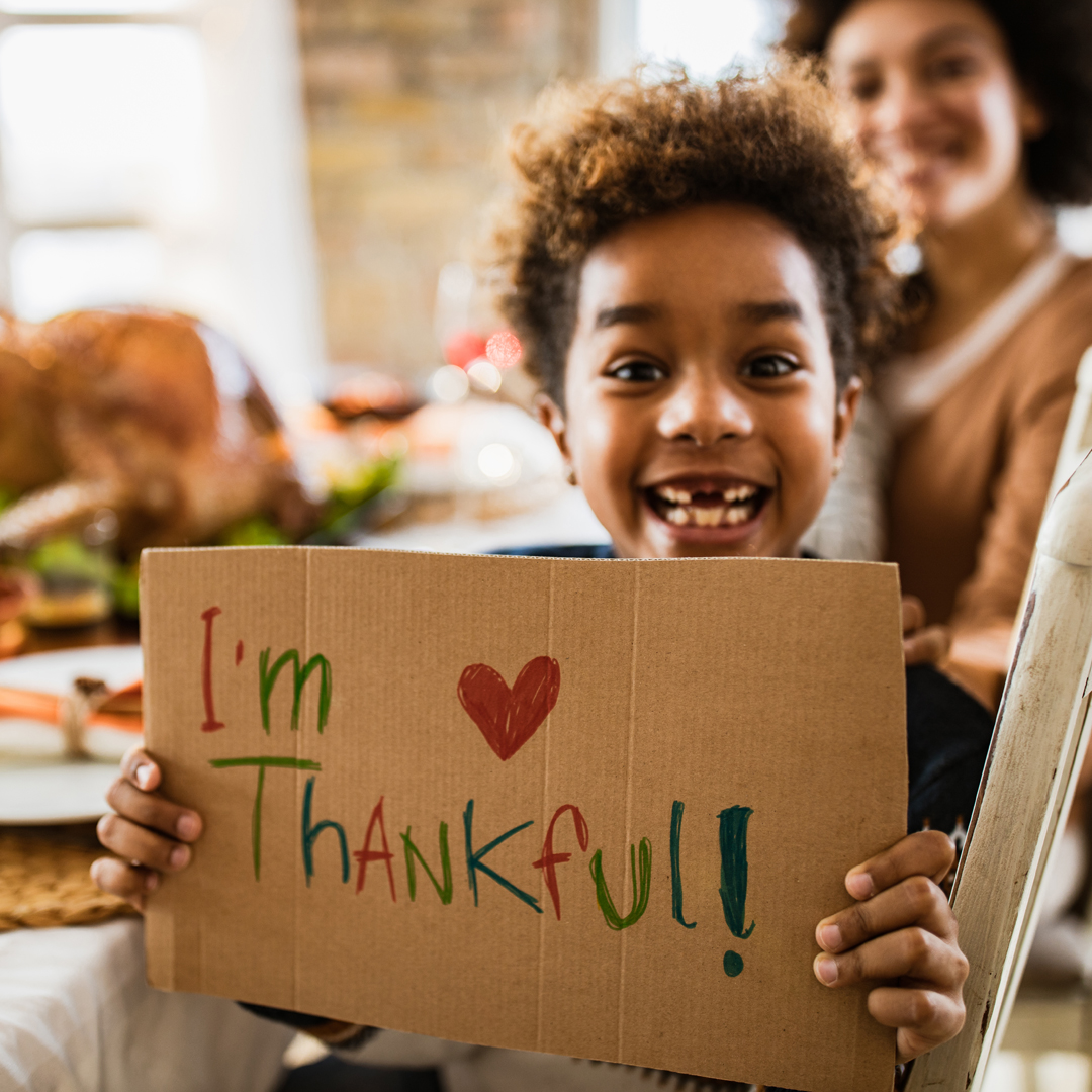 Smiling child at the Thanksgiving table holding a sign that says I’m thankful 