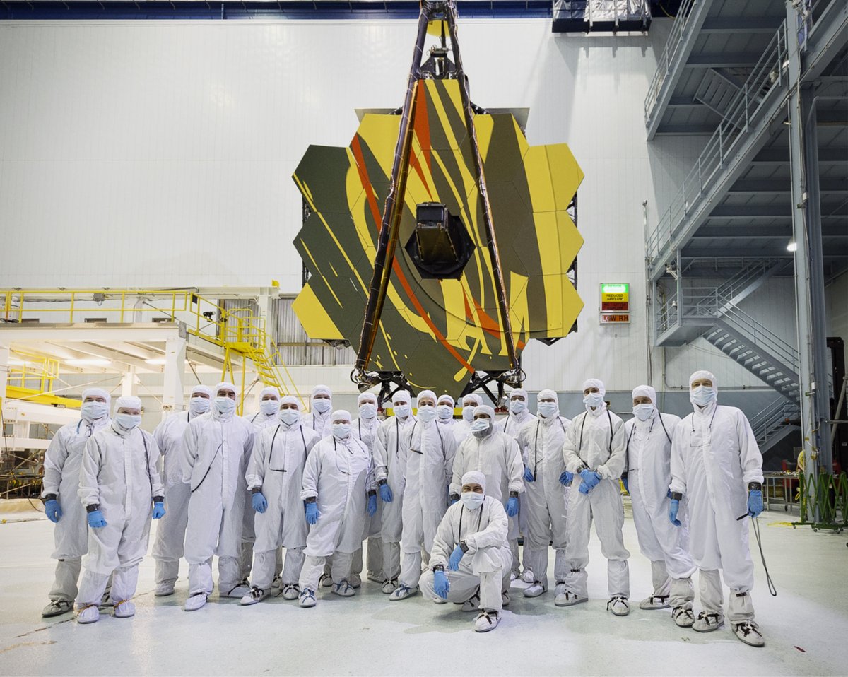 A group of technicians in cleanroom suits pose in front of the Webb Telescope's primary mirror in the NASA Goddard Spaceflight Center cleanroom.