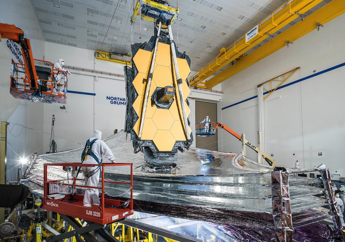 The James Webb Space Telescope's silver kite-shaped sunshield is fully deployed in the Northrop Grumman cleanroom. A single technician in a white cleanroom suit stands on an orange crane to the left of the telescope's primary mirror.
