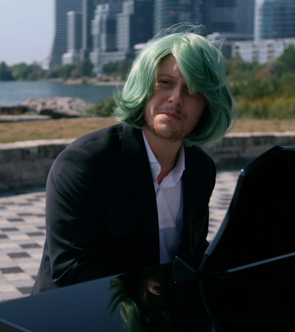 Noah Reid wearing the same mint green wig. He sits at the piano with his head tilted to his left.