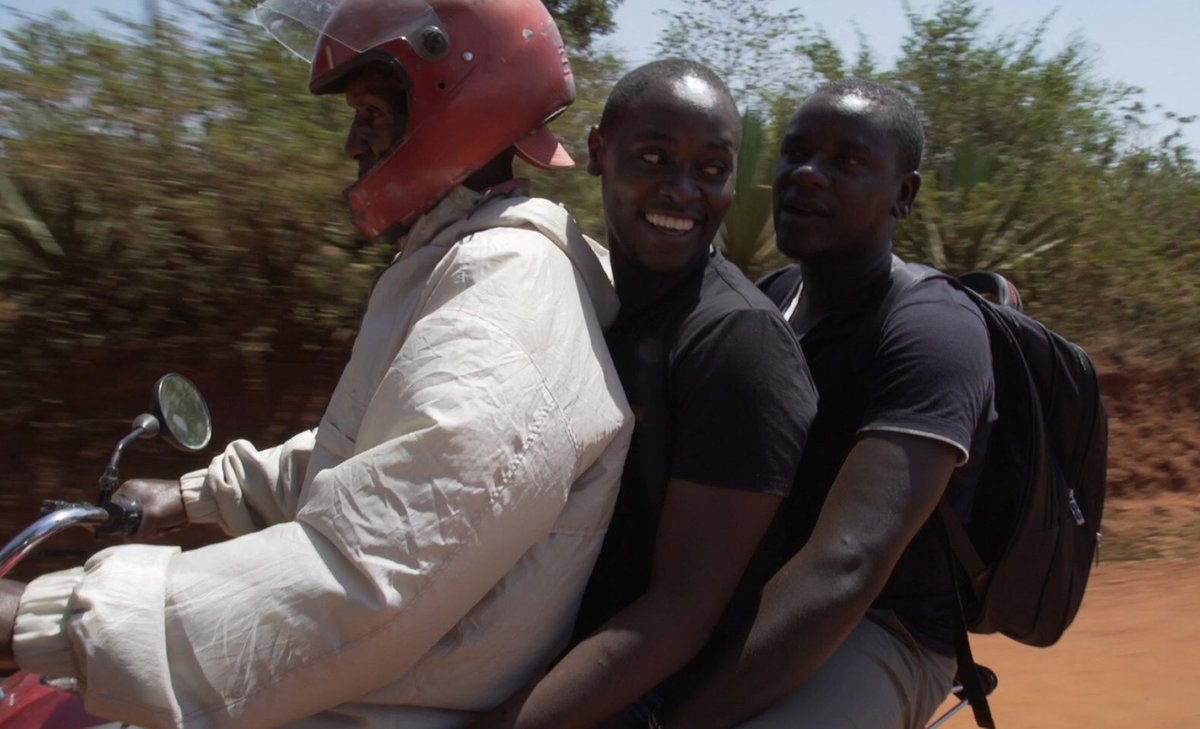 Two Kenyan men sit on the back of a moped smiling and holding tight as they drive down a road in the Kenyan countryside.