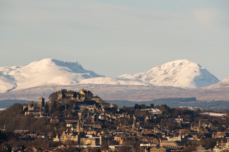 A city with a hill and stone castle on top at its centre. Snow-covered mountains can be seen in the background