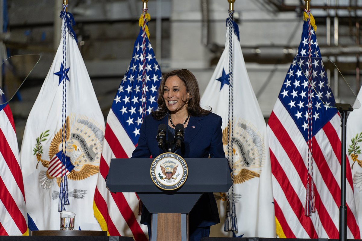 Vice President Harris speaks from a lectern that has the vice presidential seal. Flags are lined up behind her, alternating between the U.S. flag and one carrying the vice presidential seal. She's wearing a navy blue suit.