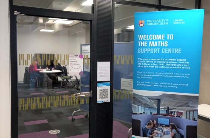 photograph of the entrance to the Maths Support Centre on the 1st floor of the Main Library. Outside the room is a blue pull up banner saying "welcome to the maths support centre" and inside the room we can see two people working. 