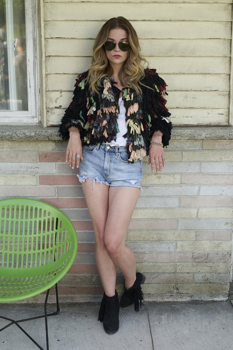 Annie Murphy on the set of Schitt’s Creek. In what’s now become an iconic outfit of Alexis’s, she leans against the wall of the motel. She wears jorts and a shaggy black cardigan that has various pastille coloured strands throughout like yellow, green and pink.