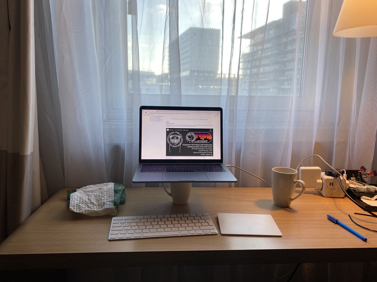 A photo of a desk in a hotel room in front of a window. Sam’s laptop is balanced on a mug, with a Bluetooth keyboard and trackpad in front of it.