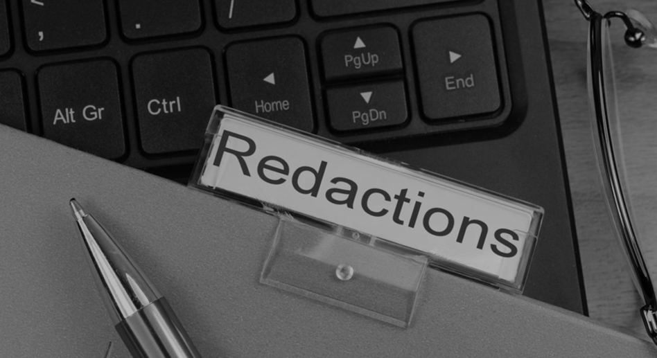 File labeled "redacted" sitting on top of a computer keyboard. The tip of a pen is laying on on file in bottom left corner.