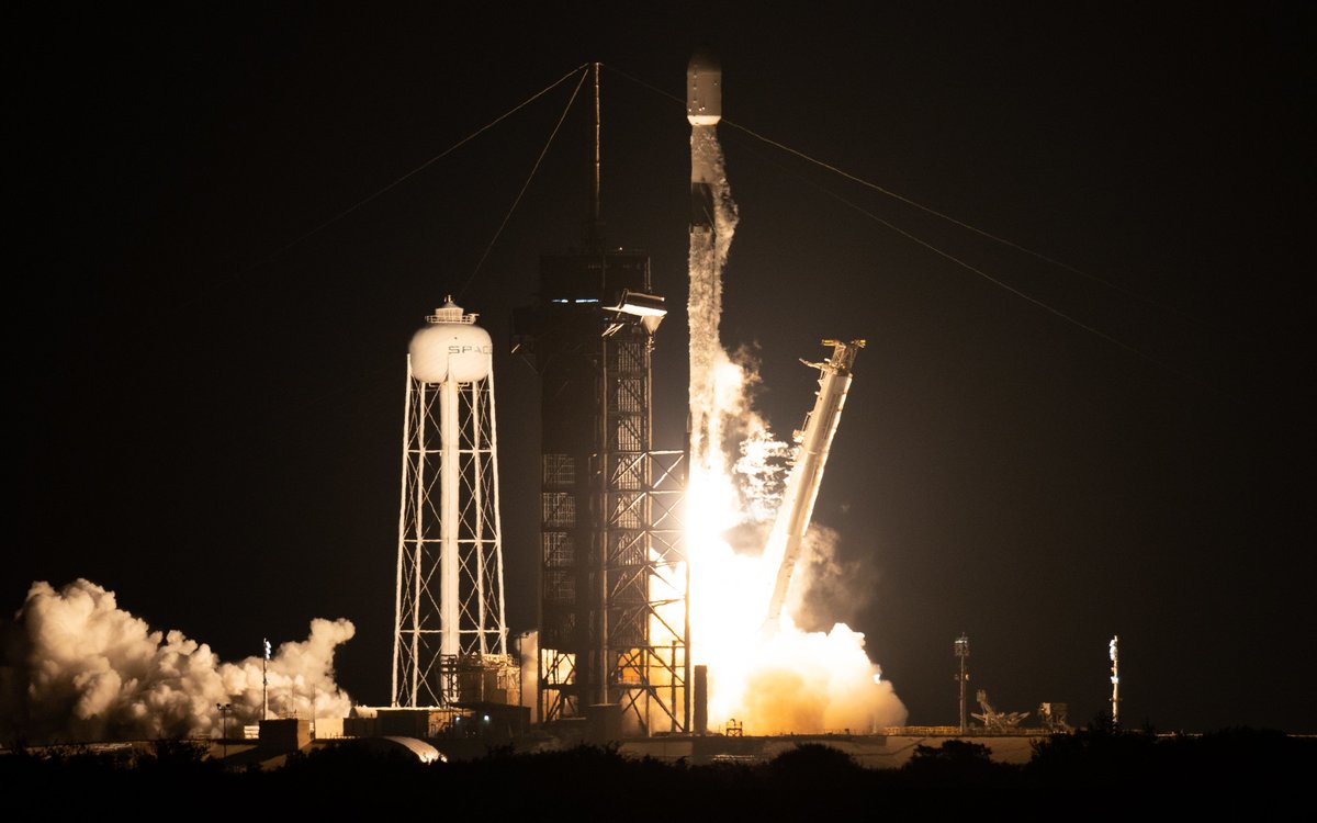 A photo of NASA's IXPE spacecraft lifting off in a SpaceX Falcon 9 rocket, a tall white cylinder with flames beneath against a pitch-black night sky