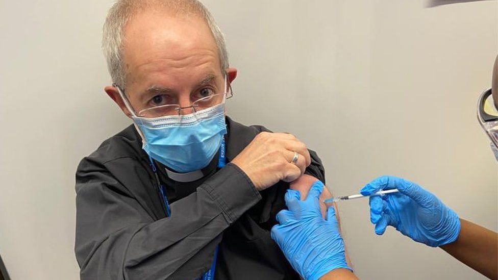 Archbishop of Canterbury shown wearing a mask, with his sleeve rolled up, being given his vaccination. 