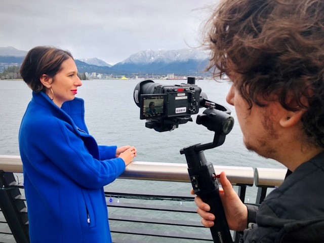 Person in a long coat looking out towards the ocean, away from a video camera which is in frame.