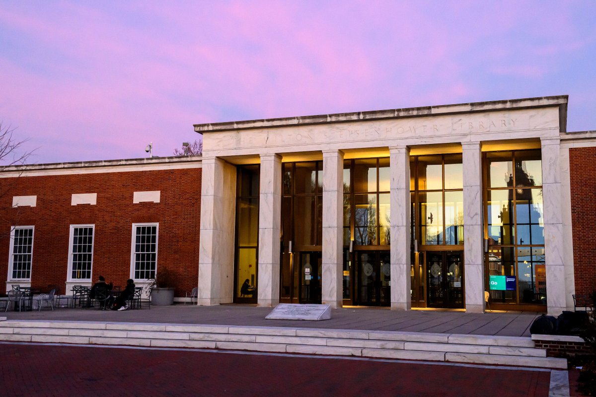 The Milton S. Eisenhower Library at sunset, with pink and purple skies above it. Reflected in the glass windows and doors of the library's entrance is the silhouette of Gilman Hall's clock tower and the glowing yellow light of the setting sun.