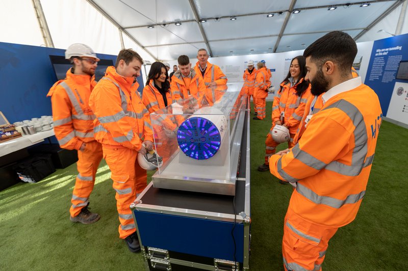Young people wearing orange protective clothing stand around a model of a tunnel boring machine in a clear case.