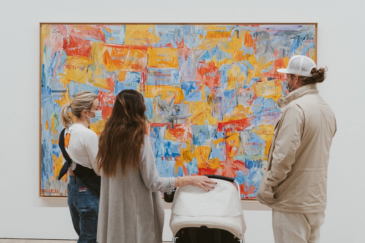 Two parents and a child in the Whitney's galleries look at Jasper Johns's painting of a flag of the United States of America, painted in sketchy brushstrokes in blue, yellow, orange, and red.